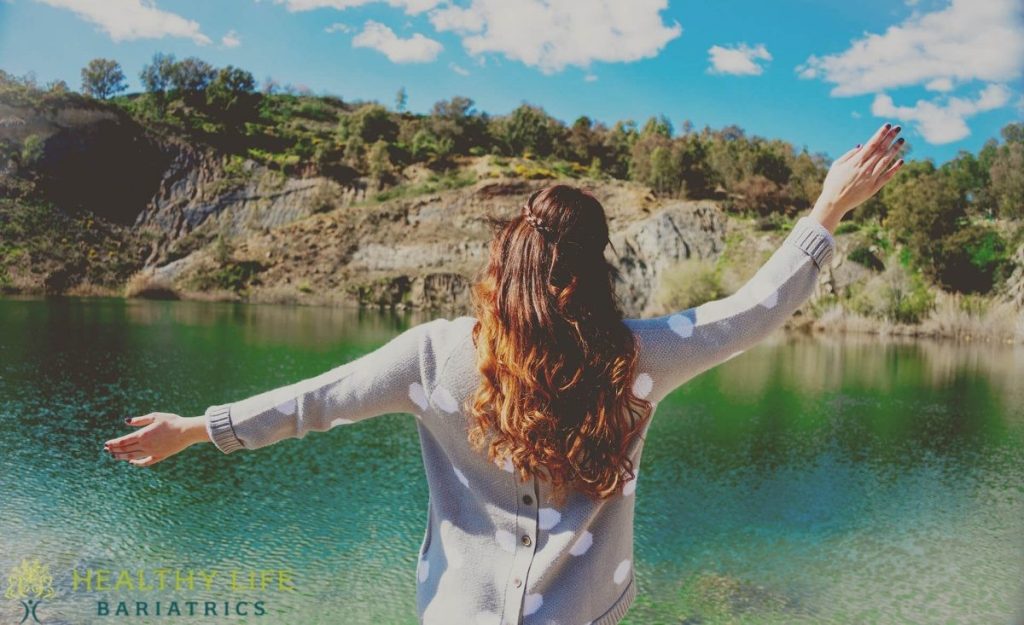 A woman standing in front of a lake with her arms outstretched.