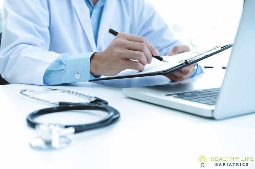 A doctor writing on a clipboard in front of a laptop.