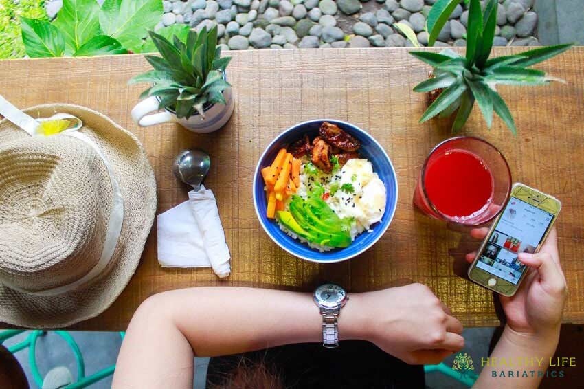 A woman is sitting at a table with a bowl of food and a cell phone.