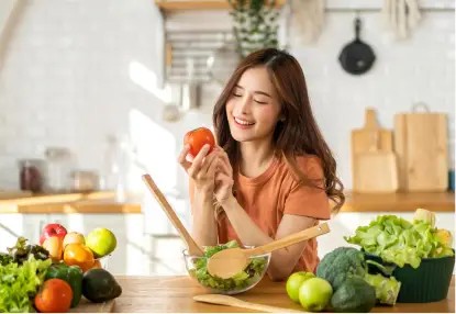 A joyful woman holding a tomato and reading a recipe in a bright kitchen filled with fresh vegetables, after her weight loss surgery in Los Angeles.