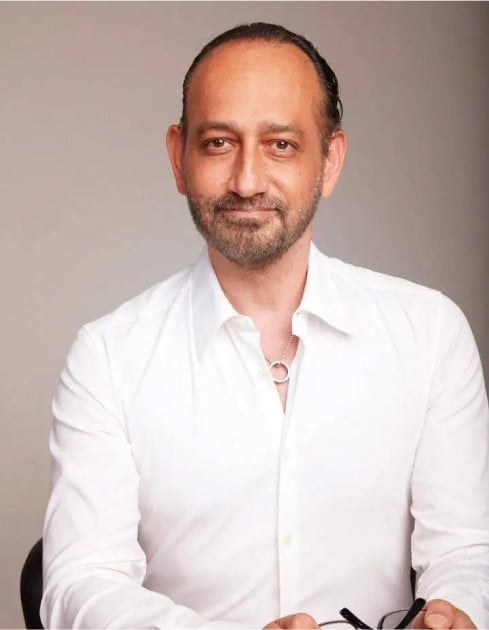 Middle-aged man with a beard, wearing a white shirt and holding glasses, sitting against a plain background in Los Angeles.