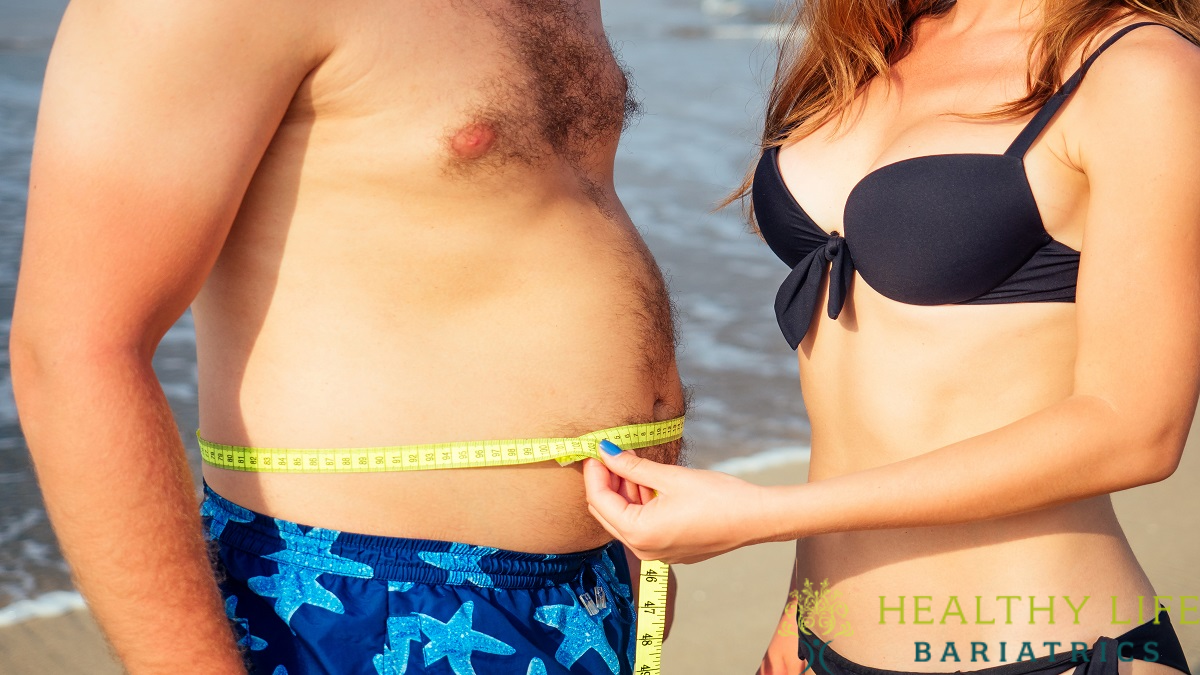 A woman in a black bikini measures a man's waist with a yellow measuring tape on a beach, likely to see if he might qualify for gastric sleeve surgery. The logo "Healthy Life Bariatrics" is visible in the bottom right corner.